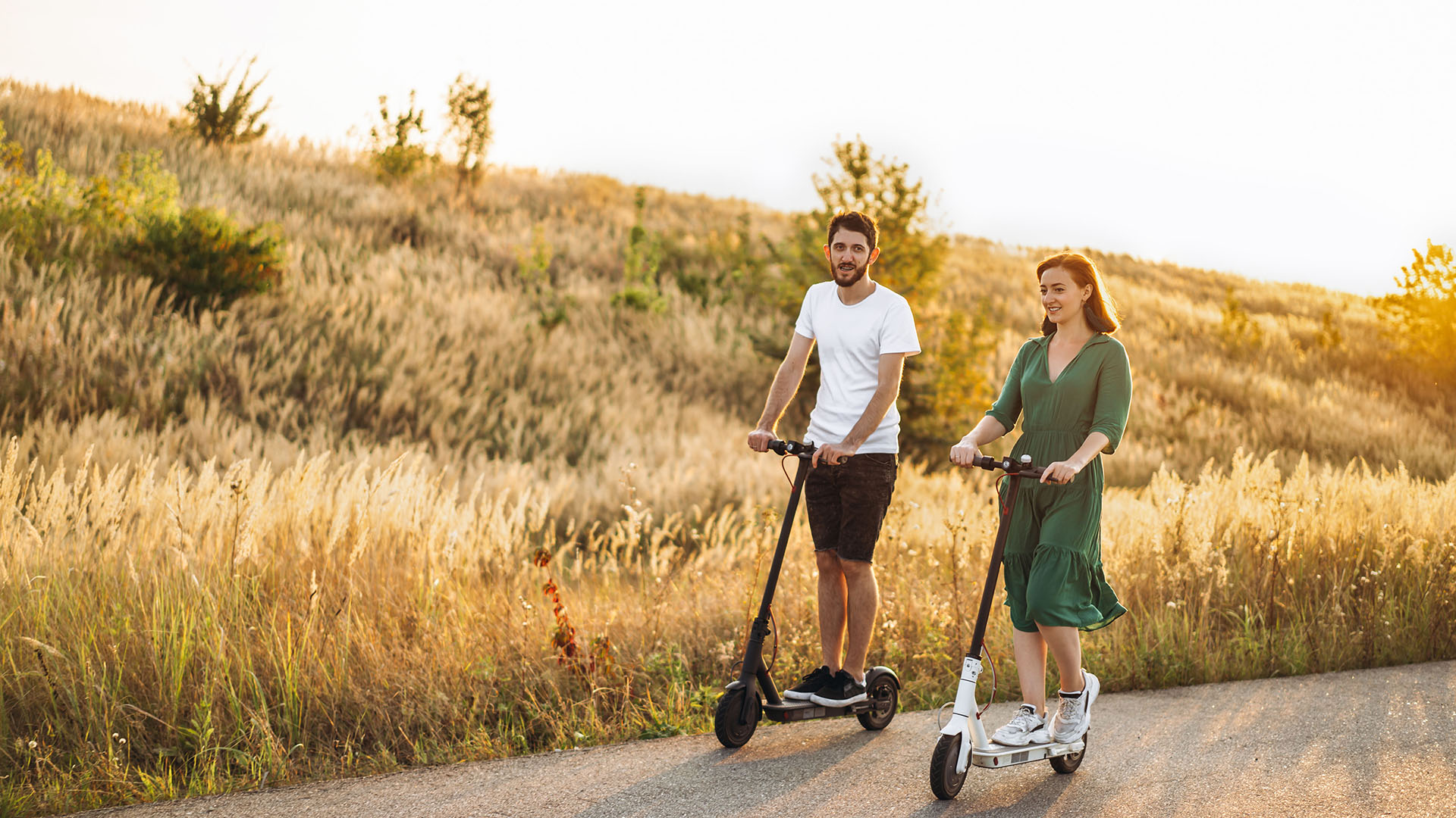 A man and woman ride electric scooters on a paved path surrounded by golden fields during sunset. 